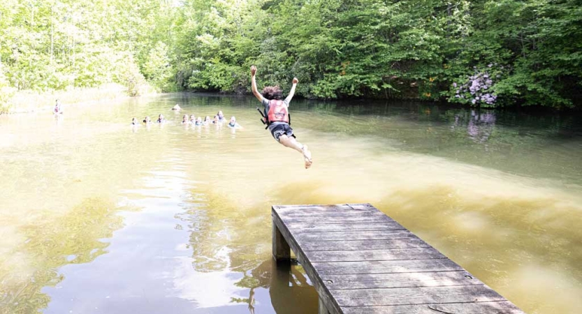 A person wearing a lifejacket is mid-air after jumping off a dock into muddy water.